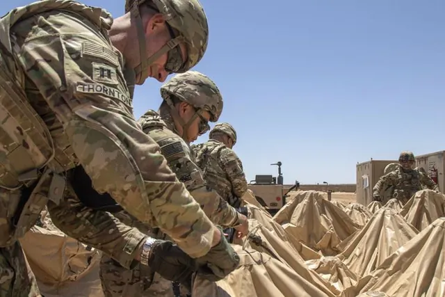 Soldiers in camouflage uniforms and helmets fold and pack tan tent canvas at a sunlit desert base