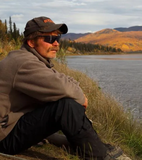 Brian Heaphy in a cap and sunglasses sitting on a grassy riverbank, with autumn hills across the water