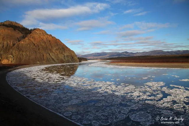 Pancake ice drifting on a wide river beside a rocky bluff, with distant mountains under a blue sky
