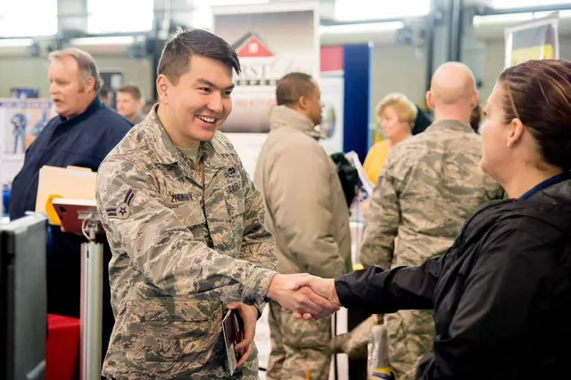 Smiling U.S. Air Force airman in camouflage shaking hands with a woman at a busy job fair, with exhibitor banners behind