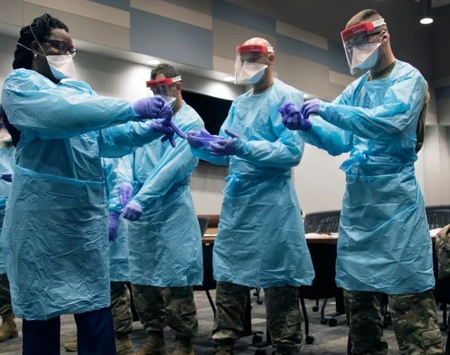 Soldiers in camouflage trousers and a woman in scrubs practice putting on blue gowns, face shields and purple gloves in a meeting room