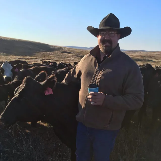 Justin Ringler in a black cowboy hat and glasses, holding a coffee cup beside a herd of black cattle on open prairie