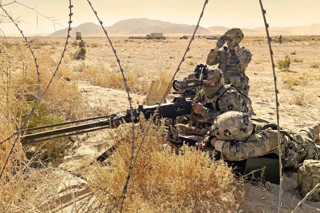 Soldiers lie behind concertina wire in dry desert brush with a heavy machine gun and sight while another soldier scans with binoculars
