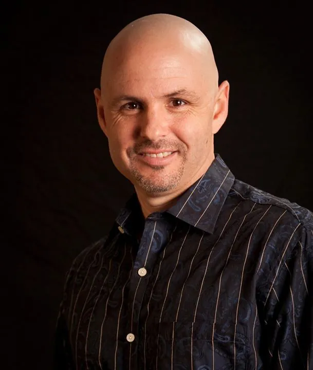 Erik Therwanger smiling in a dark striped, patterned shirt against a black background