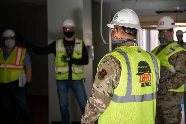 Masked Guard members in hard hats and safety vests tour an unfinished building with construction staff