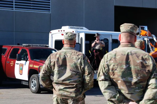 Two Guard soldiers, seen from behind, listen to a Minnesota State Patrol trooper speaking from a bus doorway beside a State Patrol truck