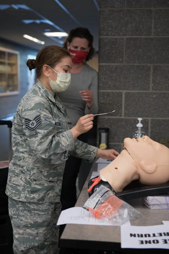 Masked airman practices a nasal swab on a training mannequin as a woman in a red mask looks on