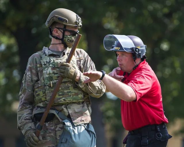 Guard soldier in a helmet and body armor holding a baton takes instruction from a trainer in a red shirt and riot helmet