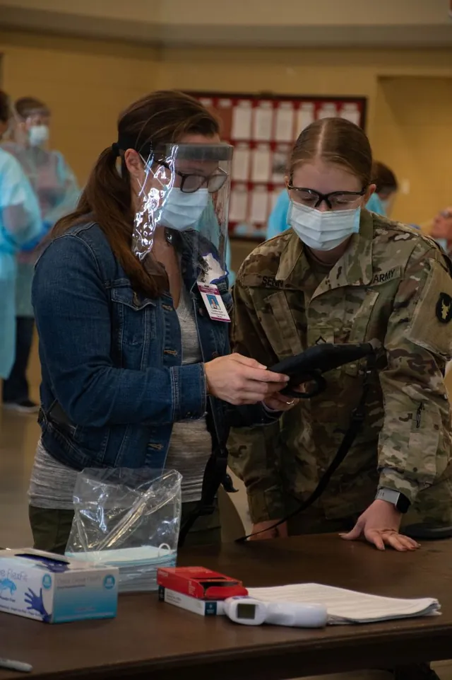 Masked soldier and a civilian in a face shield look at a handheld device at a COVID-19 testing table with gloves and a thermometer