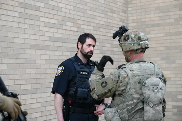 Helmeted Guard soldier gestures while talking with a uniformed police officer beside a brick wall