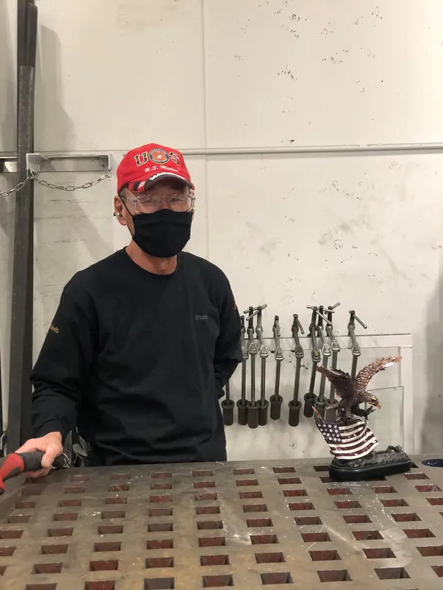 John Kish in a red U.S. Marine cap, safety glasses and face mask stands at a Nystrom welding table with the eagle trophy beside him