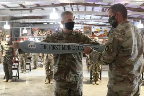 Two masked soldiers stretch out a 2nd Battalion, 135th Infantry streamer reading The First Minnesota in front of seated troops