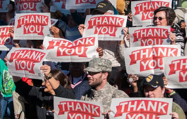 Crowd of spectators and veterans holding up red and white Thank You signs, a man in an Army camouflage cap in front.