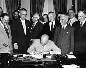 Black and white photo of President Eisenhower seated at a desk signing a bill as seven men in suits stand behind him.