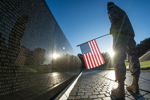 Uniformed soldier holding a small U.S. flag beside the black granite wall of the Vietnam Veterans Memorial as the sun rises.