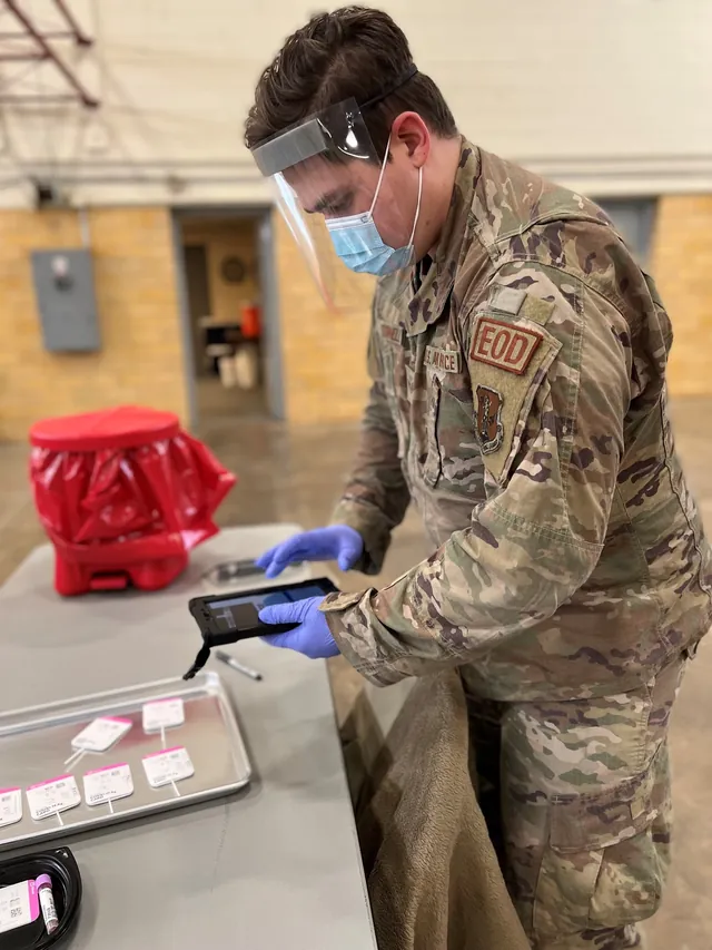 Airman in camouflage, face shield and mask checking a tablet beside a tray of COVID-19 test cards at a community testing site.