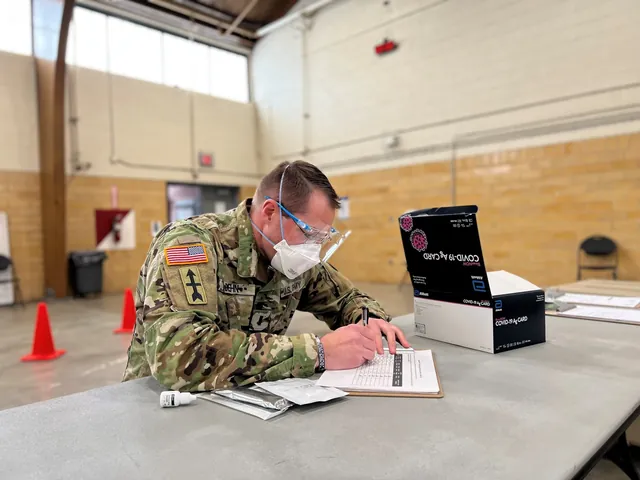 Soldier in camouflage, safety glasses and N95 mask filling in a form at a table beside a box of COVID-19 antigen test cards.