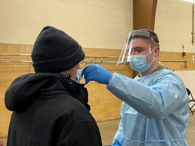 Tester in a face shield, mask and blue gown taking a nasal swab from a man in a black winter hat and coat.