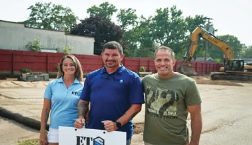 Three people holding an ETS sign on a cleared construction lot with an excavator behind them