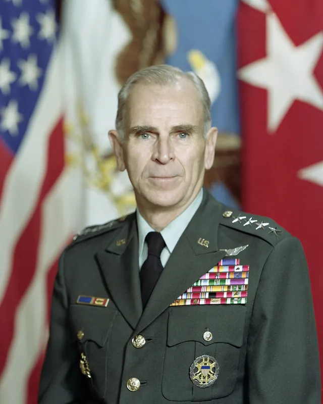 General John W. Vessey Jr. in Army green uniform with four-star shoulder insignia and rows of ribbons, standing before flags