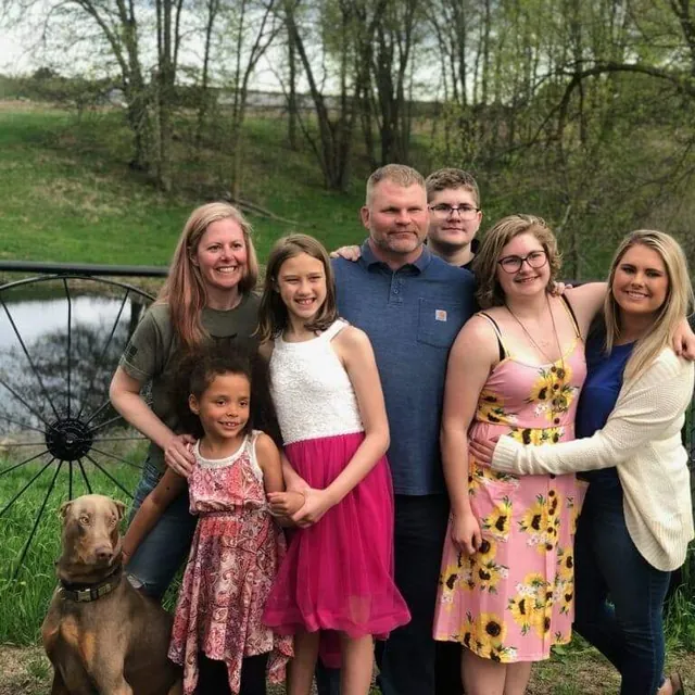 Jason Rehn with six family members and a brown Doberman on a lawn, with a pond, an old wagon wheel and spring trees behind them