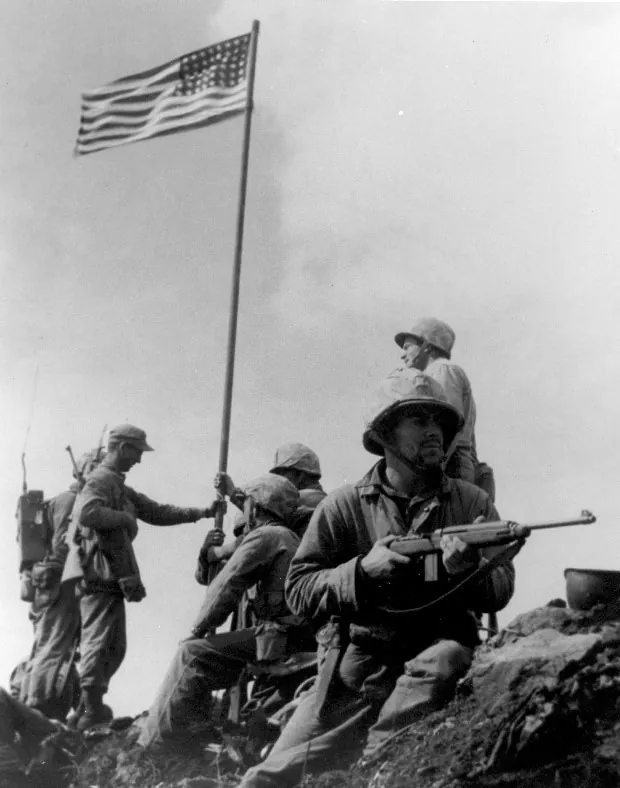 Marines raising an American flag on a long pole atop Mount Suribachi while a Marine with a carbine keeps watch in the foreground