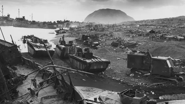 Wrecked landing craft, amphibious tractors and equipment on Iwo Jima’s black sand beach, with Mount Suribachi and landing ships behind