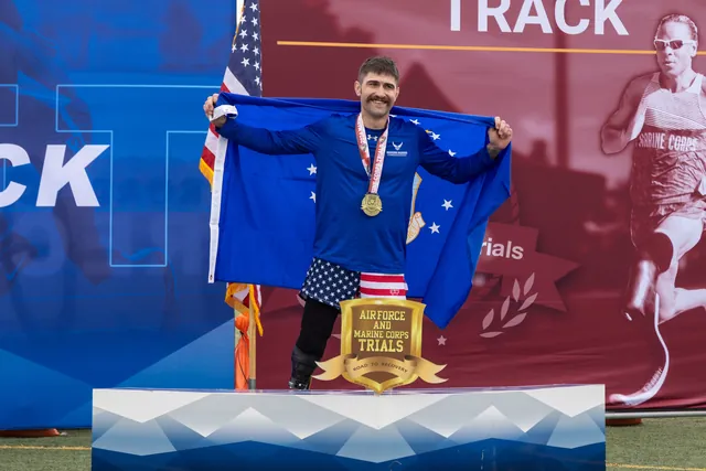 Brandon Sullivan, a medal around his neck, holds a blue flag with white stars on the podium at the Air Force and Marine Corps Trials