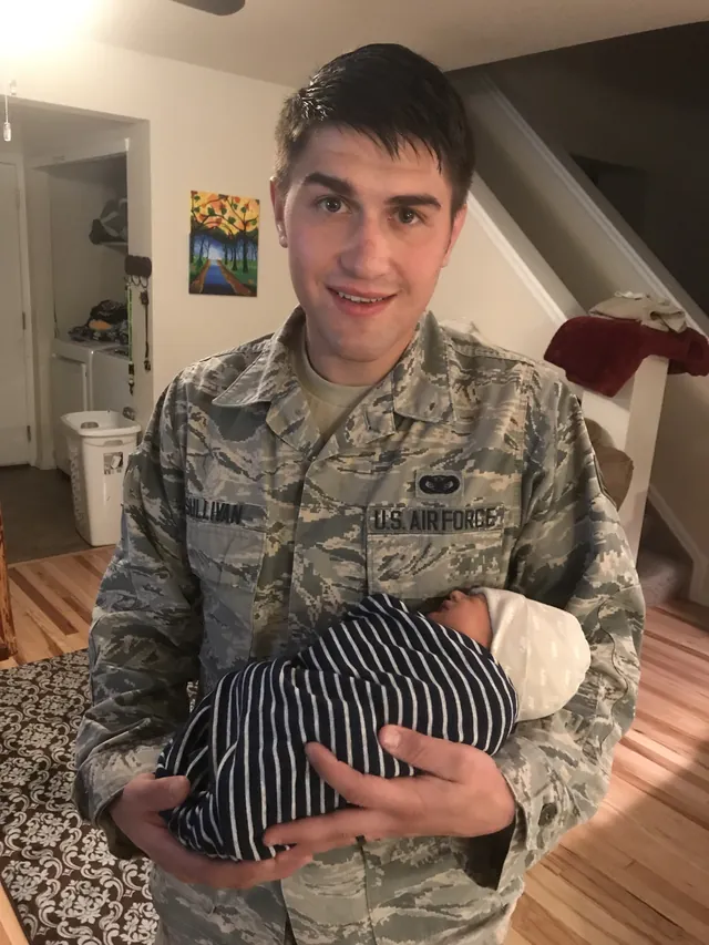 Brandon Sullivan in his Air Force uniform, name tape reading Sullivan, cradles a newborn baby wrapped in a striped blanket