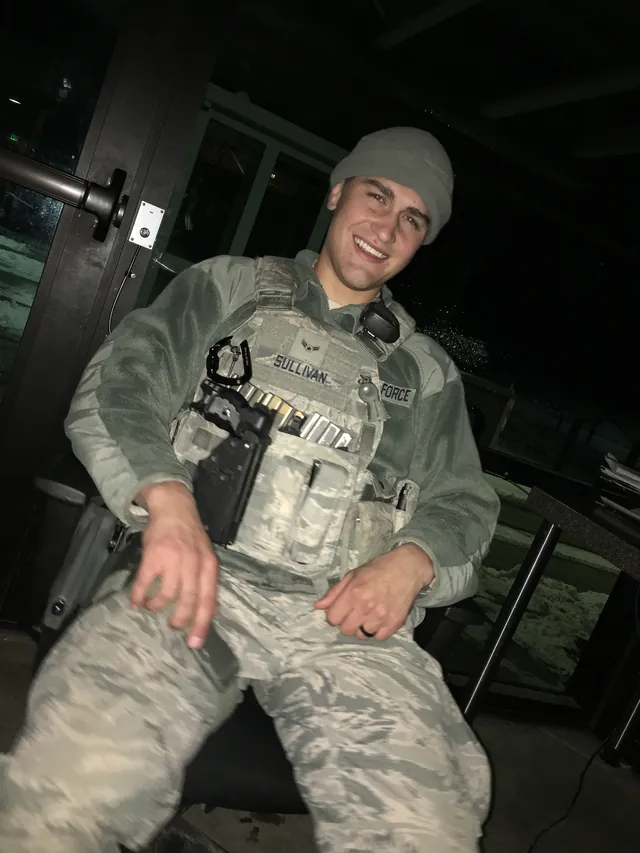 Brandon Sullivan in Air Force camouflage, a knit cap and a tactical vest with his name tape, sitting and smiling at night