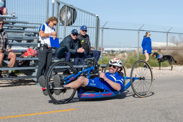 Brandon Sullivan, in a helmet and sunglasses, races a blue hand cycle past spectators seated in the bleachers