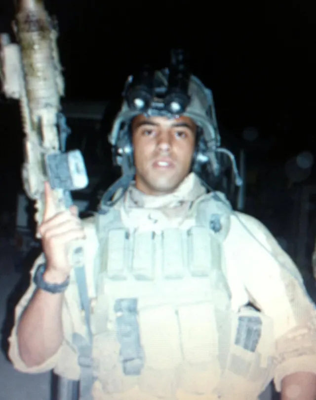 John Mitchell at night in desert combat gear and a helmet with night-vision goggles, holding his rifle upright