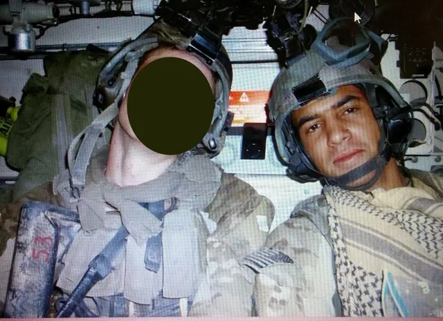 John Mitchell in a combat helmet and checked scarf, seated beside a fellow soldier whose face is blacked out