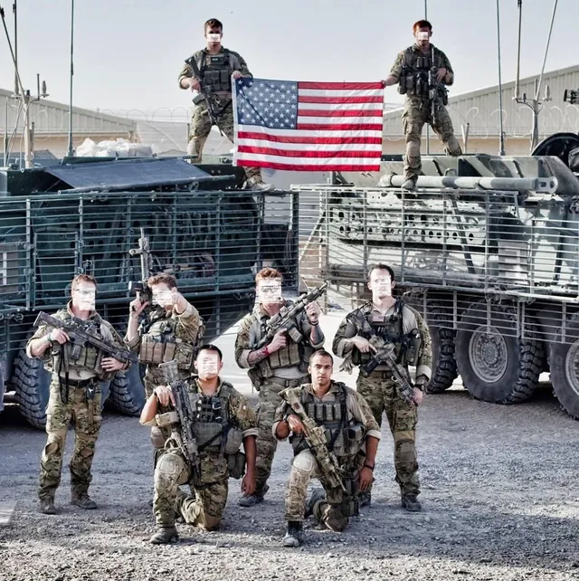 Soldiers in combat gear, faces blurred, pose with rifles in front of armored vehicles as two hold up a U.S. flag; John Mitchell kneels in front