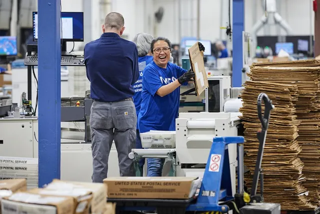 Smiling IWCO worker in a blue company T-shirt lifts a flat carton on a production floor beside stacks of cardboard and a pallet jack