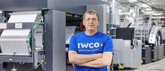 IWCO employee in a blue IWCO T-shirt and safety glasses stands with arms crossed in front of large printing presses on the plant floor