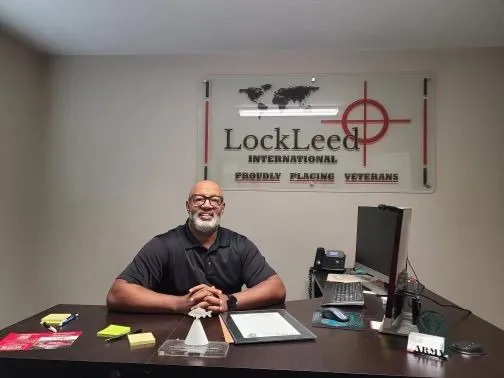 James Walker smiles at a desk with a phone and computer, under a wall sign reading LockLeed International, Proudly Placing Veterans