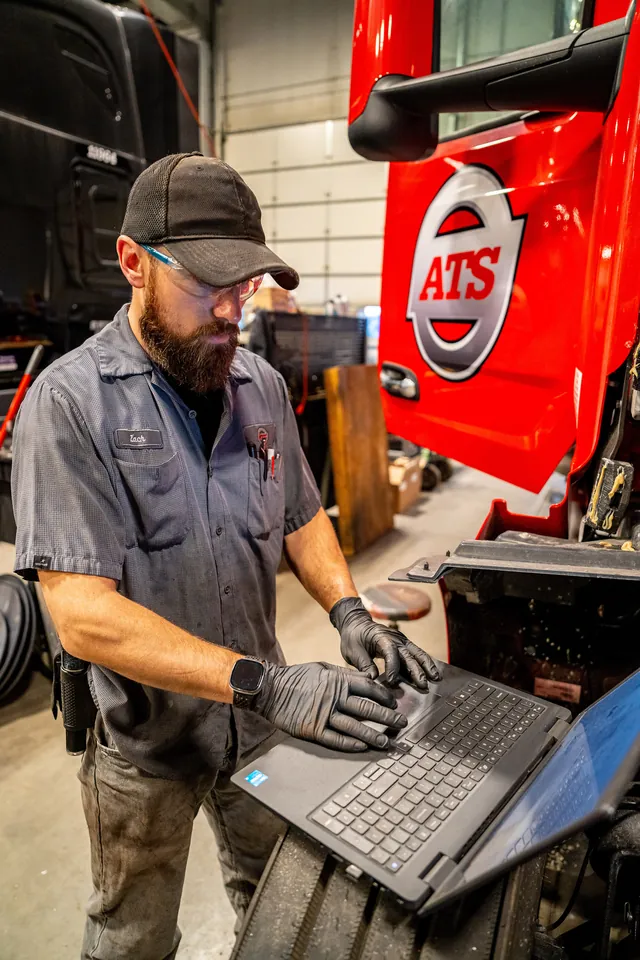 Bearded ATS technician in a cap, safety glasses and gloves works on a laptop beside the open cab of a red ATS truck in a shop