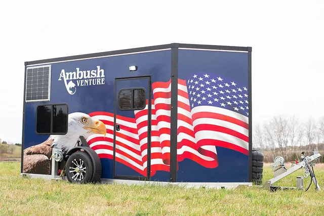 Ambush Venture ice fishing house on a wheeled trailer, wrapped in an American flag and bald eagle graphic, parked on a grassy field