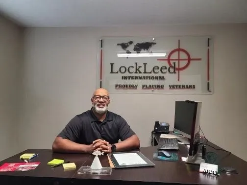 Jim Walker smiling at a desk in front of a wall sign reading LockLeed International, Proudly Placing Veterans, with an Army nameplate on the desk