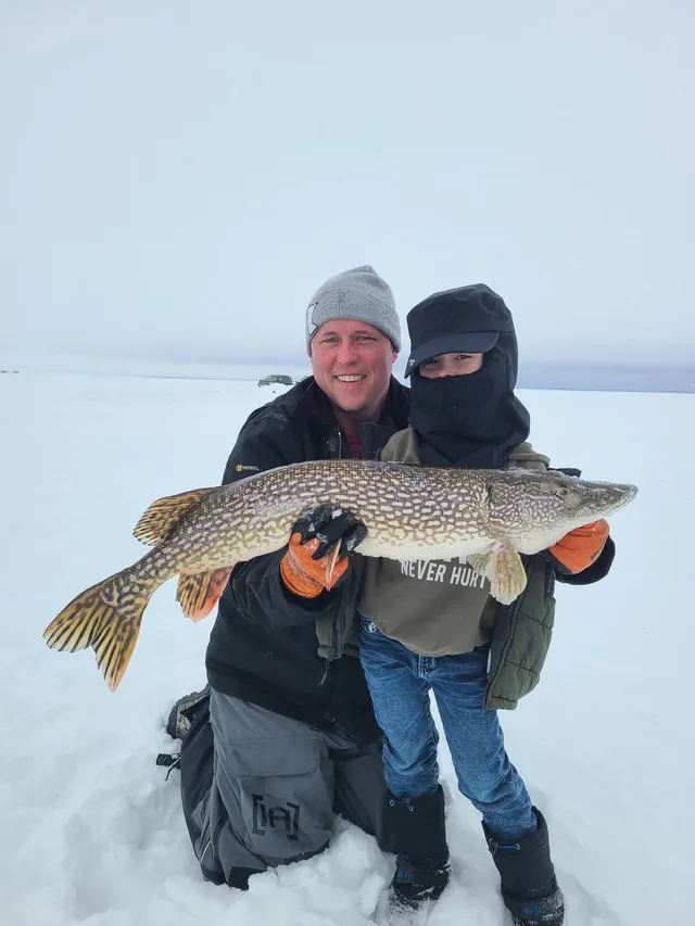Smiling man in a gray knit cap kneels on a snowy frozen lake beside a bundled-up child, the two holding up a large northern pike