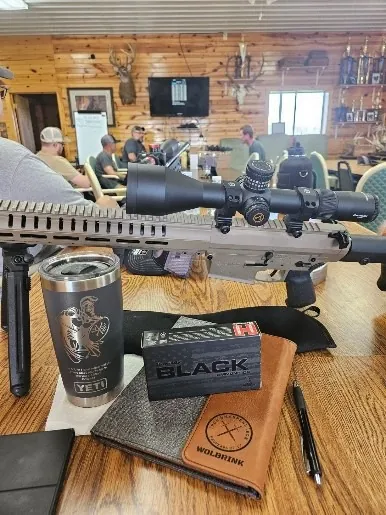 Scoped rifle on a table beside an engraved YETI tumbler, a box of ammunition and a leather folio engraved Wolbrink, with people seated behind