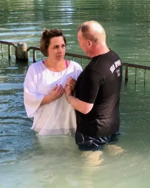 Man in a black T-shirt baptizing a woman in a white robe, both standing waist-deep in green river water beside a railing